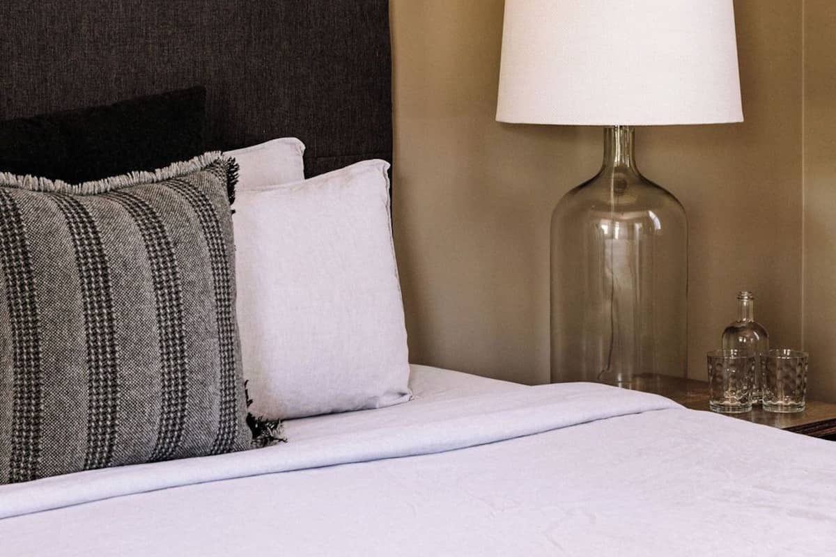 A neatly made bed with white linens, a dark headboard, and two pillows. A clear glass table lamp with a white lampshade is beside the bed.