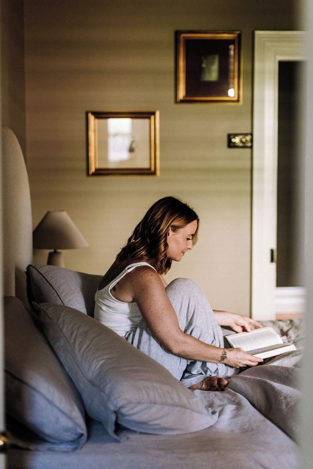 Una persona sentada en una cama leyendo un libro en una habitación con una lámpara y cuadros en la pared.