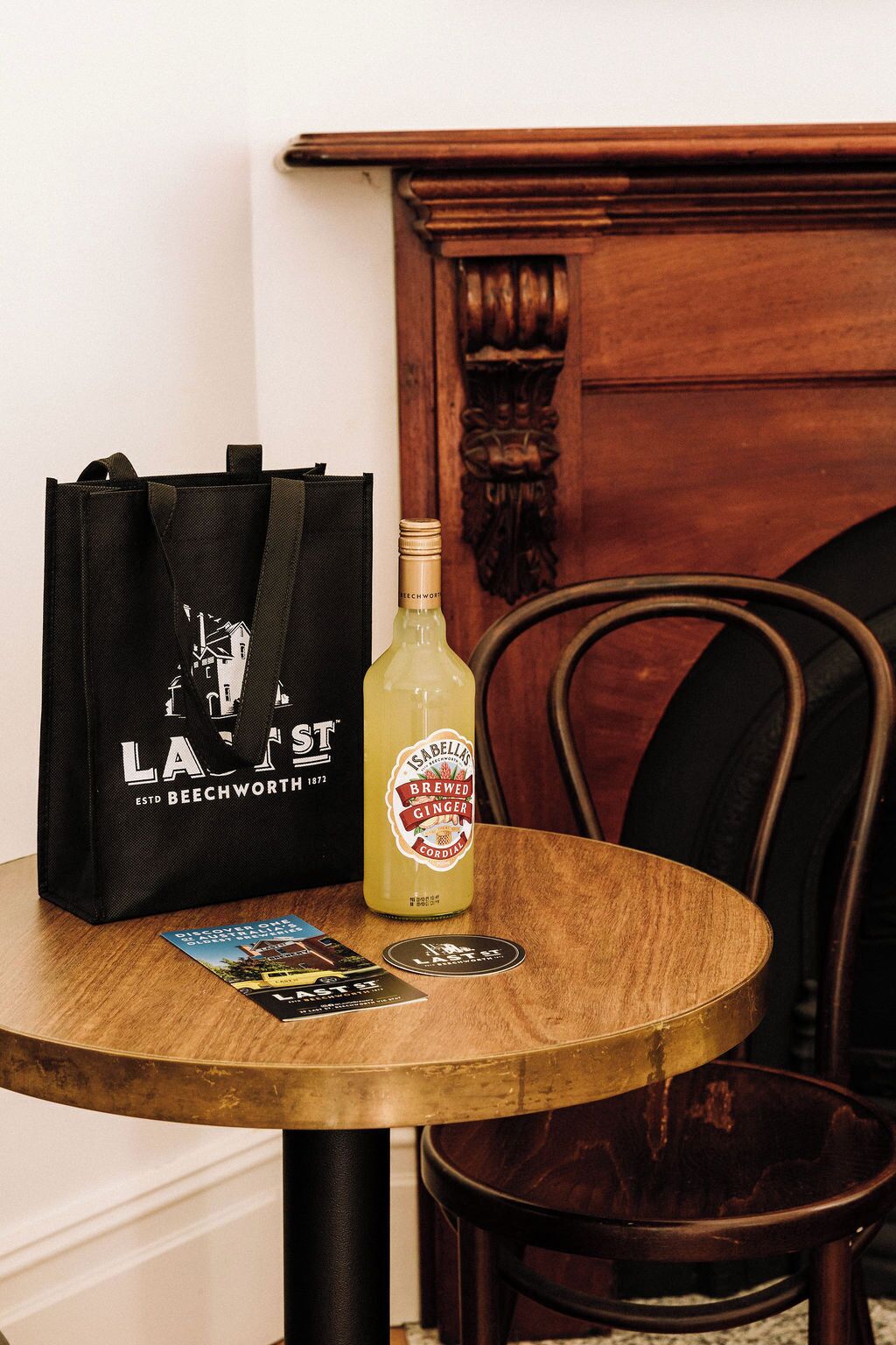 Een ronde houten tafel met een zwarte tas, een fles Isabelli's Brewed Ginger Cordial en twee ansichtkaarten.