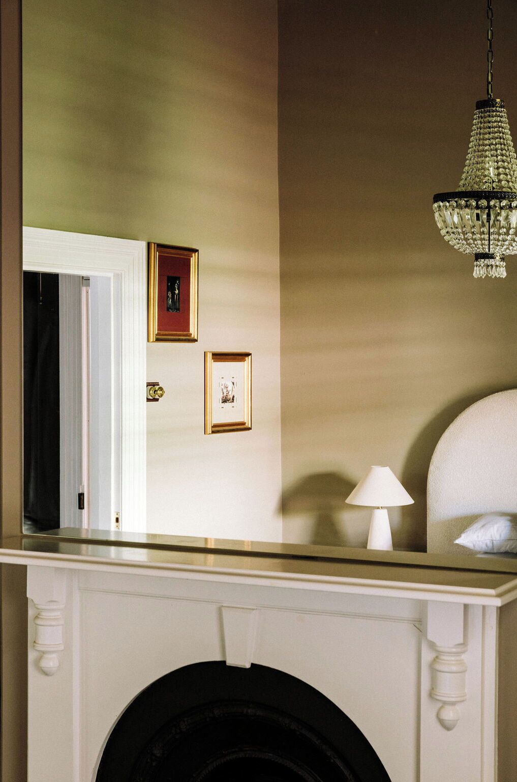 A room with a fireplace, a mirror reflecting a door and two framed pictures, and a chandelier.
