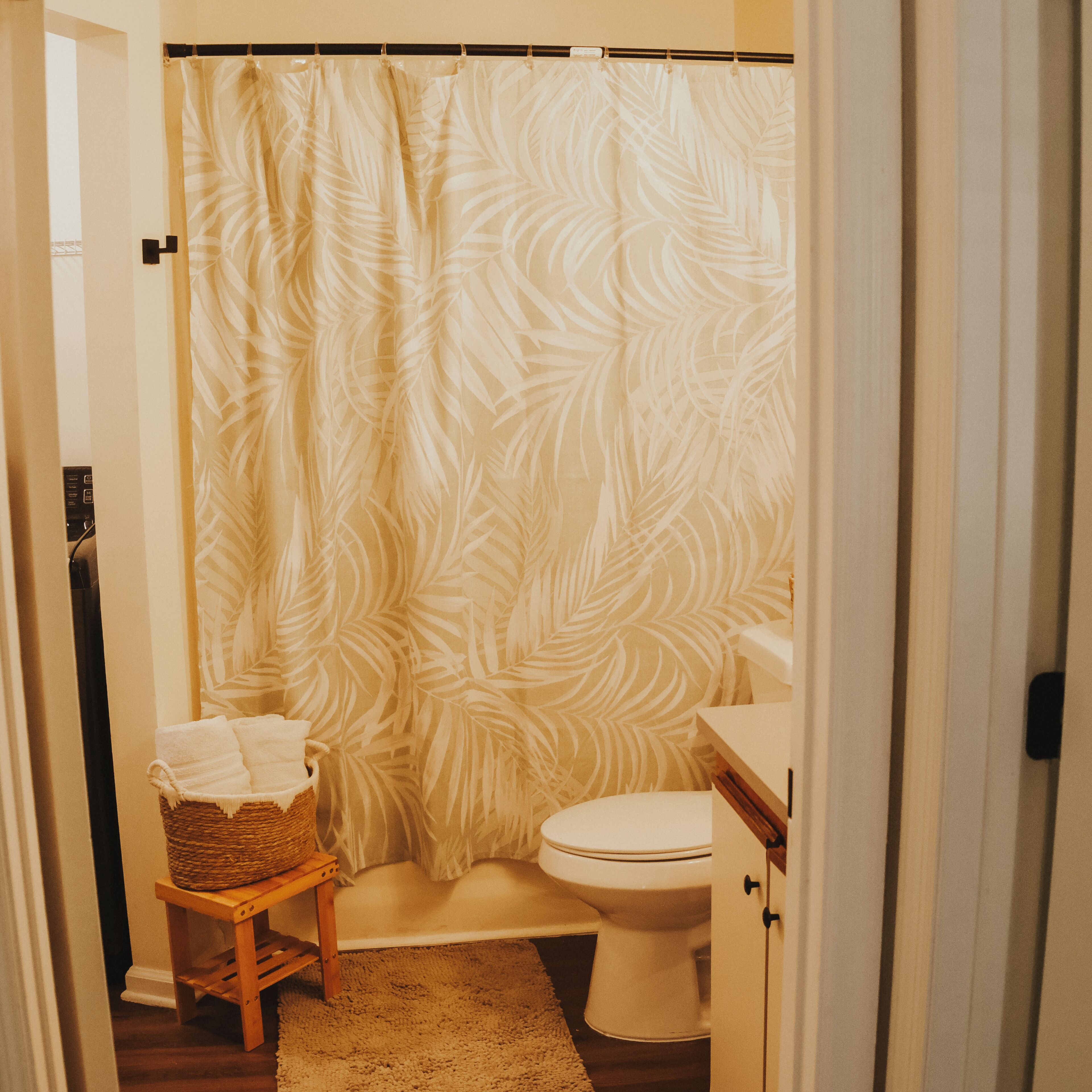 Un baño con cortina de ducha, inodoro, un taburete de madera pequeño con una cesta y un lavabo blanco.