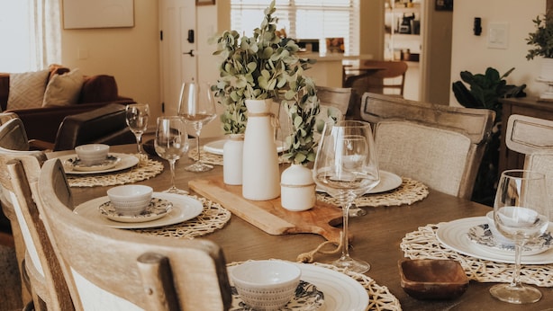 A dining table set with white plates, bowls, and wine glasses, surrounded by wooden chairs.