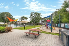 A picnic table in a park with playground equipment and a fence in the background.