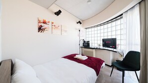 Ein modernes Hotelzimmer mit gewölbter Decke, einem Bett mit weißen Bettwäsche, einer roten Tagesdecke, einem Schreibtisch mit Computer und einem Flachbildfernseher, der 'NETFLIX' zeigt, sowie einem Fenster mit Vorhängen.