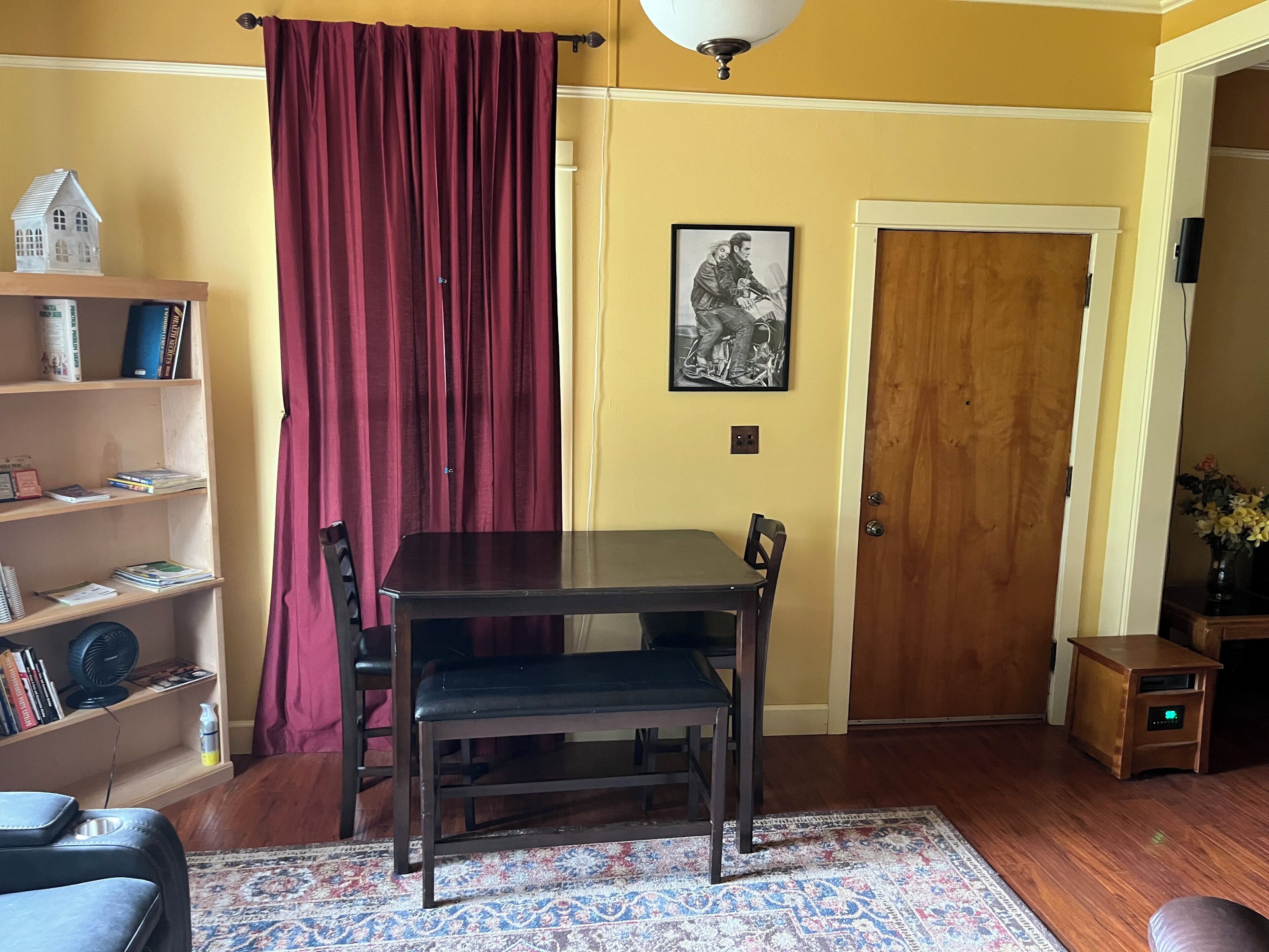 Un comedor con una mesa y sillas de madera, una estantería, una cortina roja, un cuadro enmarcado en la pared y una puerta de madera.