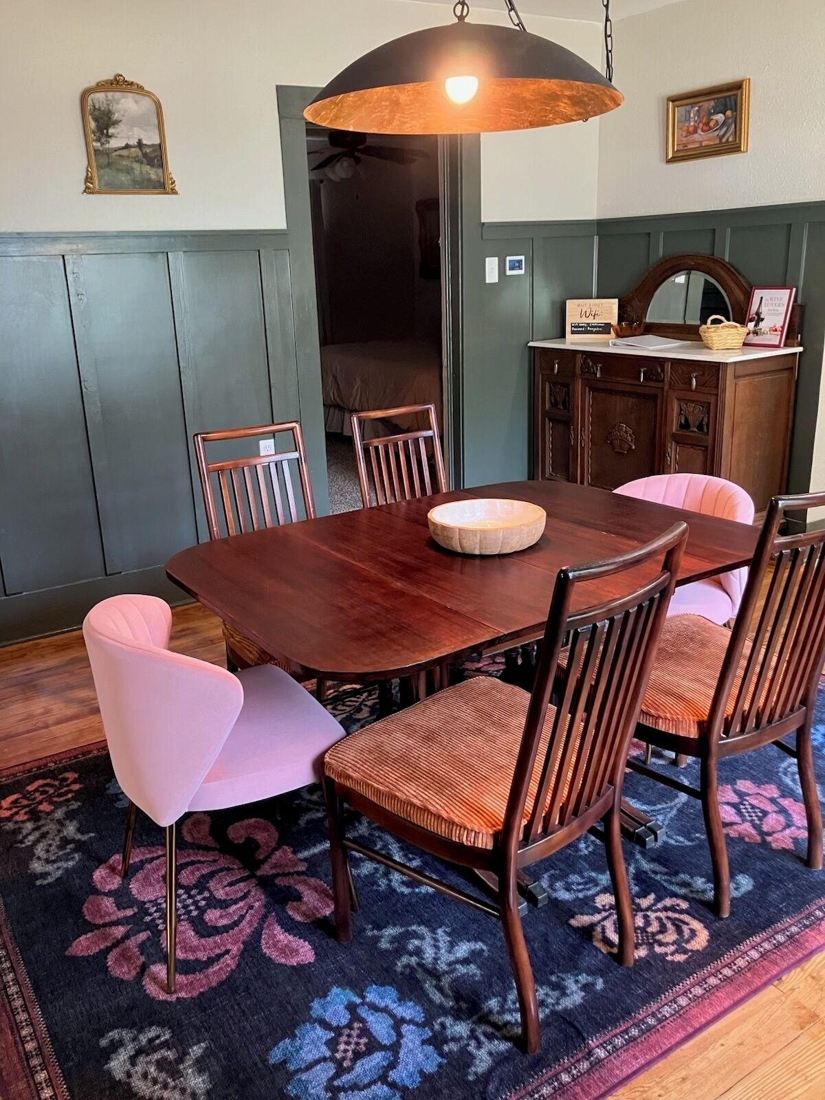 A dining area with a wooden table, six chairs, a large pendant light, a patterned rug, and a cabinet with decorative items.