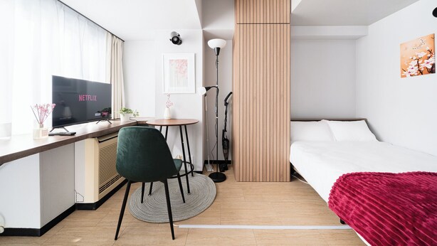 A compact hotel room with a bed, a desk with a TV displaying 'NETFLIX', a small table with a plant, a green chair, a floor lamp, and a vertical wooden panel. - Excellent access to Shibuya area! Residence with cherry blossom motif [307]. (Meguro-ku)