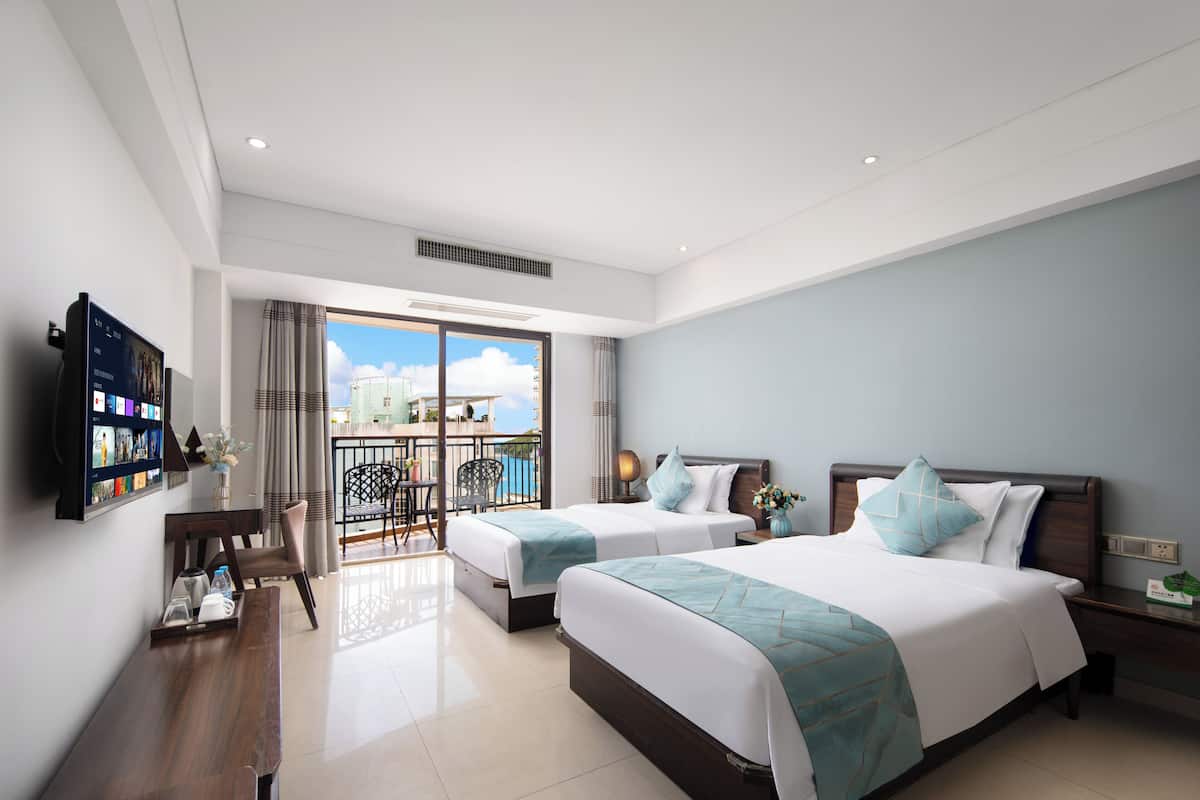 A modern hotel room with two beds, a TV, a desk, and a balcony with a view of the city.