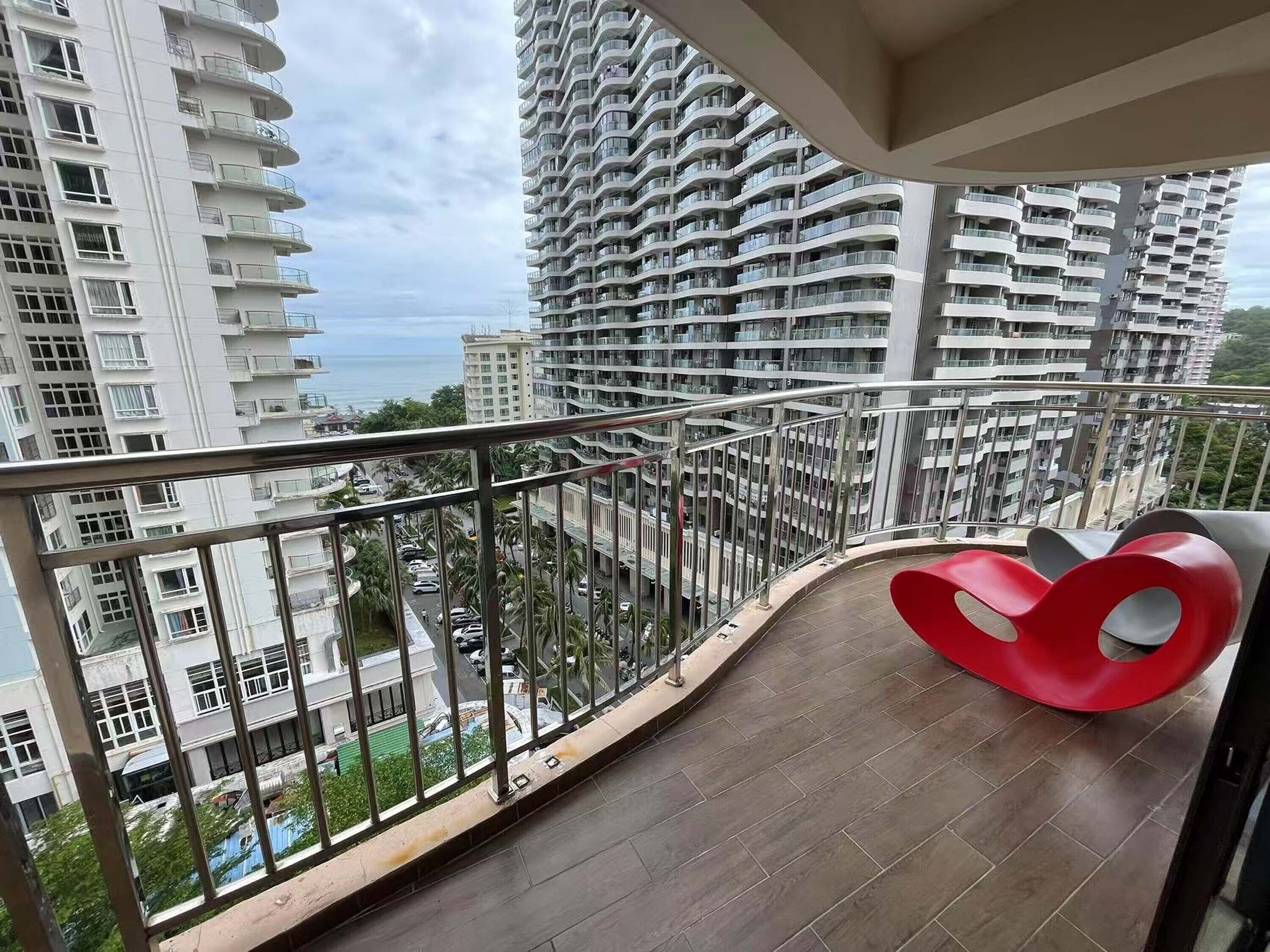 A balcony with a curved railing, a red chair, and a view of high-rise buildings and a body of water.