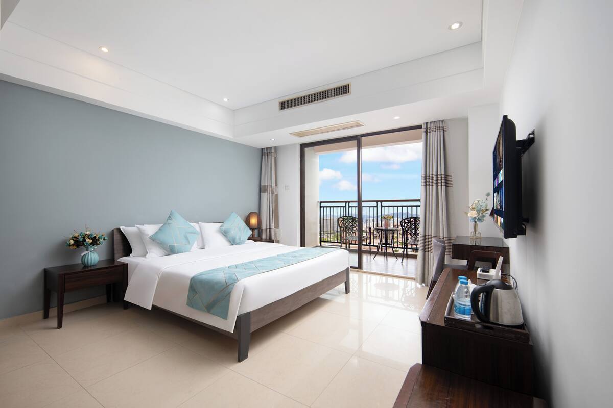 A modern hotel room with a large bed, a small desk, a TV, and a balcony with a view.