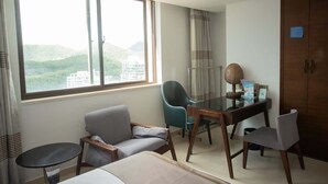 A hotel room with a bed, a desk, a chair, a small table, and a large window offering a view of a cityscape. - Sanya Xintai Ocean View Hotel (Sanya)