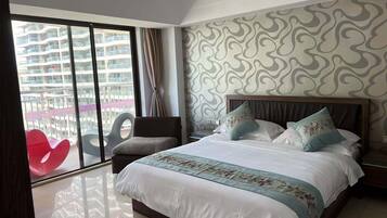 Un cuarto de hotel con una cama grande, una mesita de noche, una silla y vista a un edificio a través de un ventanal amplio.