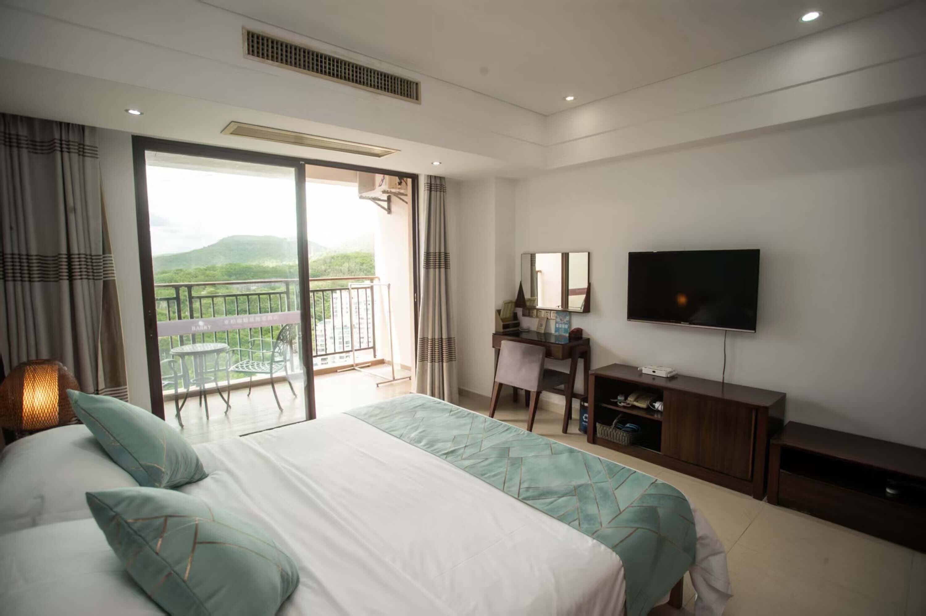 A hotel room with a large bed, a desk with a computer, a TV, and a balcony with a view of the hills.