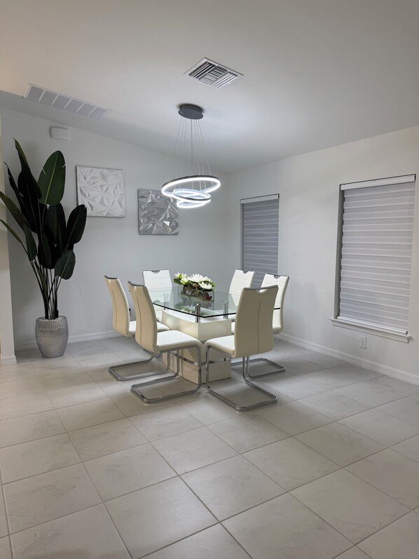 A modern dining area with a glass-top table, white chairs, a potted plant, and geometric wall art. - Family home (Cape Coral)
