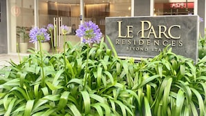 A sign for Le Parc Residences with a modern building facade and a well-maintained garden with green foliage and purple flowers.