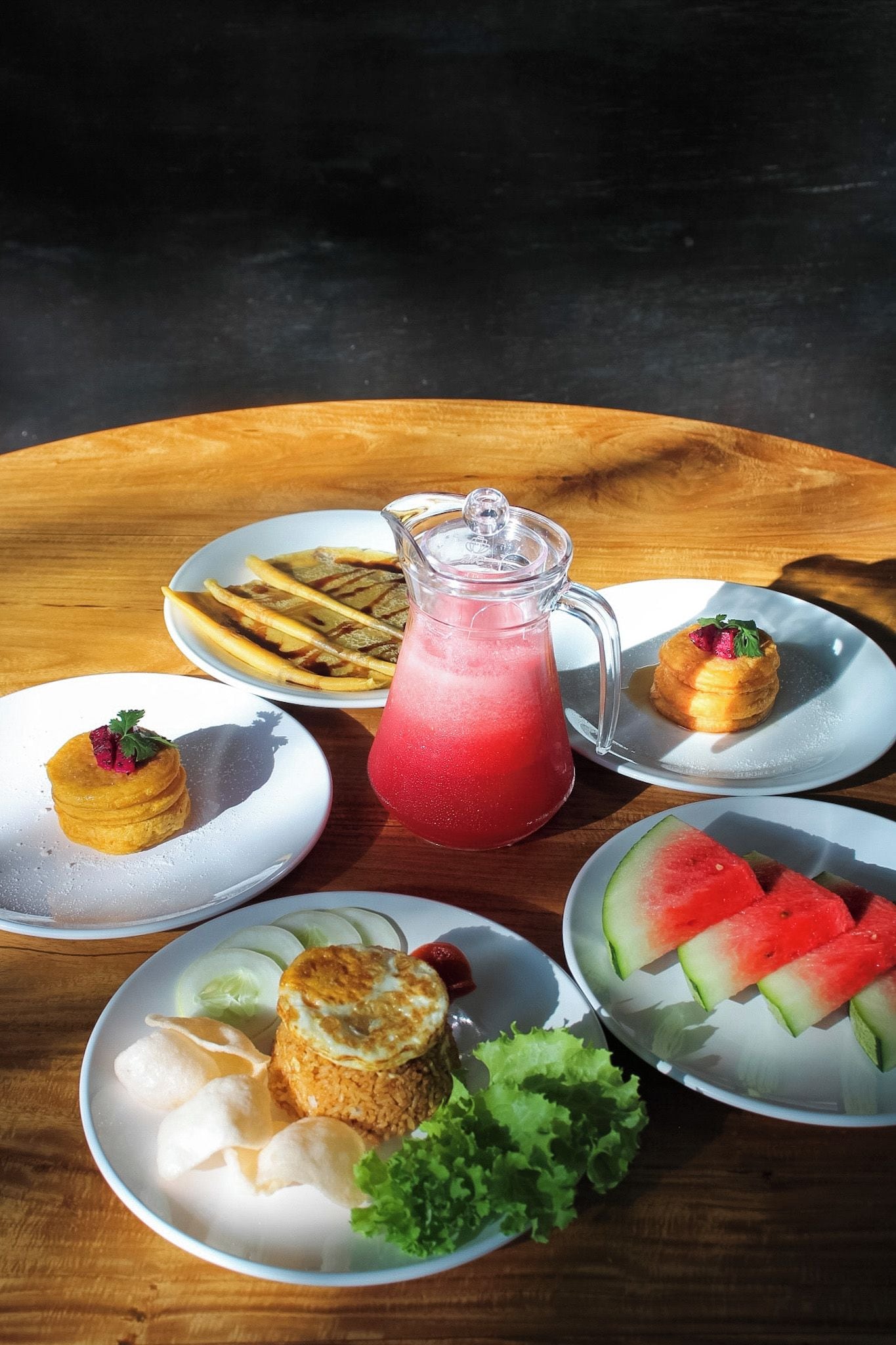 Una mesa redonda de madera con diversos alimentos, como panqueques, gofres, un jarro de bebida roja y rebanadas de sandía.