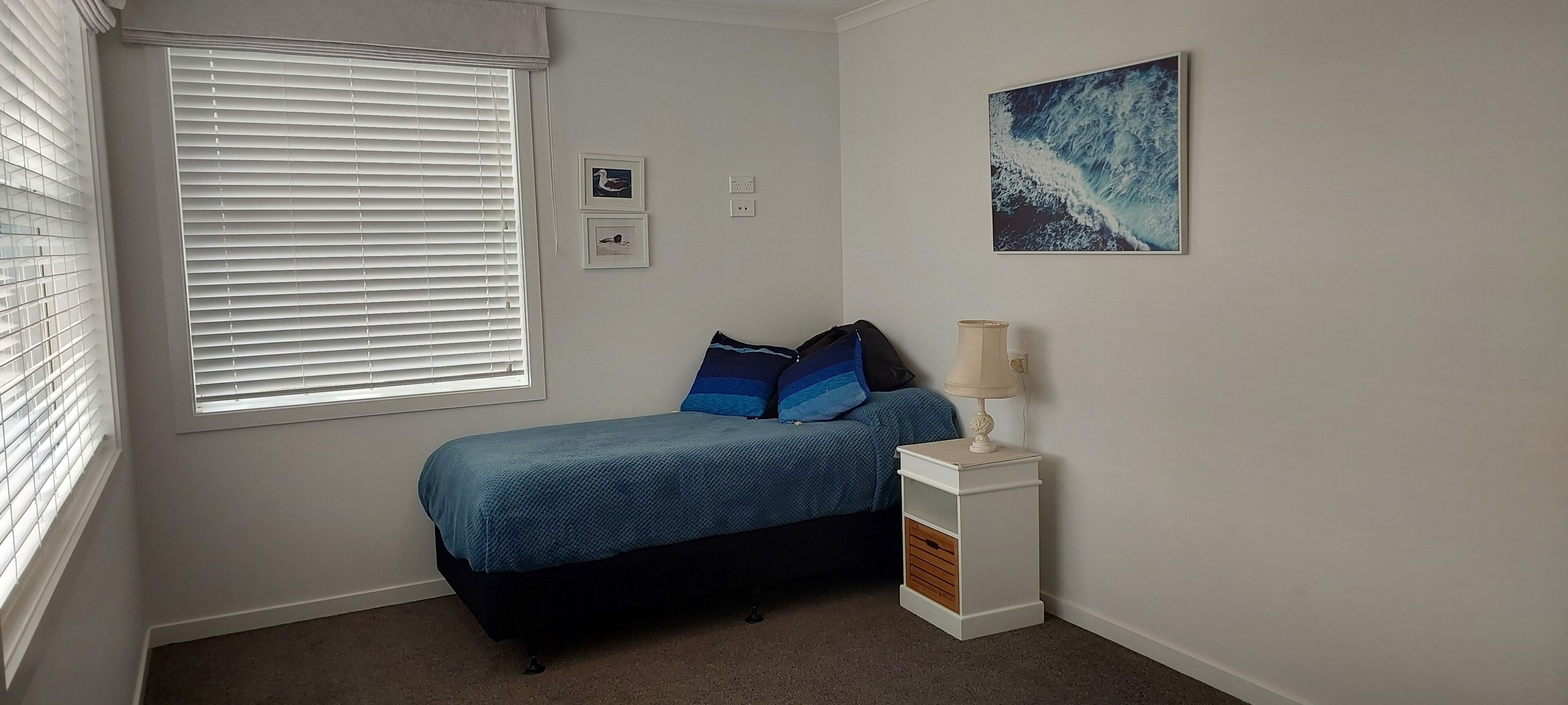 Ein Schlafzimmer mit einem Bett, einem Nachttisch, einer Lampe, einem Fenster mit Jalousien und zwei gerahmten Bildern an der Wand.