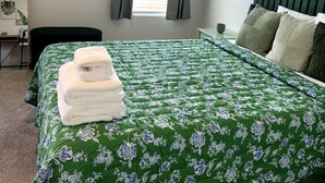 Een netjes opgemaakt bed met een bloemenbedekking in groen en wit, een stapel witte handdoeken en een groen hoofdeinde.