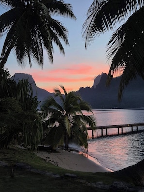 Escena de playa con palmeras, un muelle de madera y montañas al fondo.