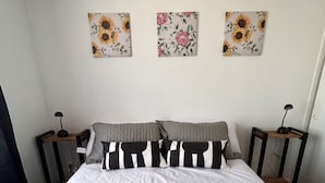 A neatly made bed with a gray quilt, black and white striped pillows, and a white pillow. There are three wall-mounted floral paintings above the bed. A white tray with a rolled towel, two white cups, and a black coffee maker is placed on the bed. - Centrally located apartment, fully equipped, with beautiful terrace, steps from the Metro. (Santiago)