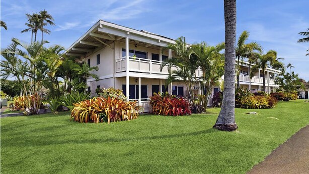 Un hotel de dos pisos con fachada blanca, balcones y jardines tropicales con palmeras y plantas de colores.