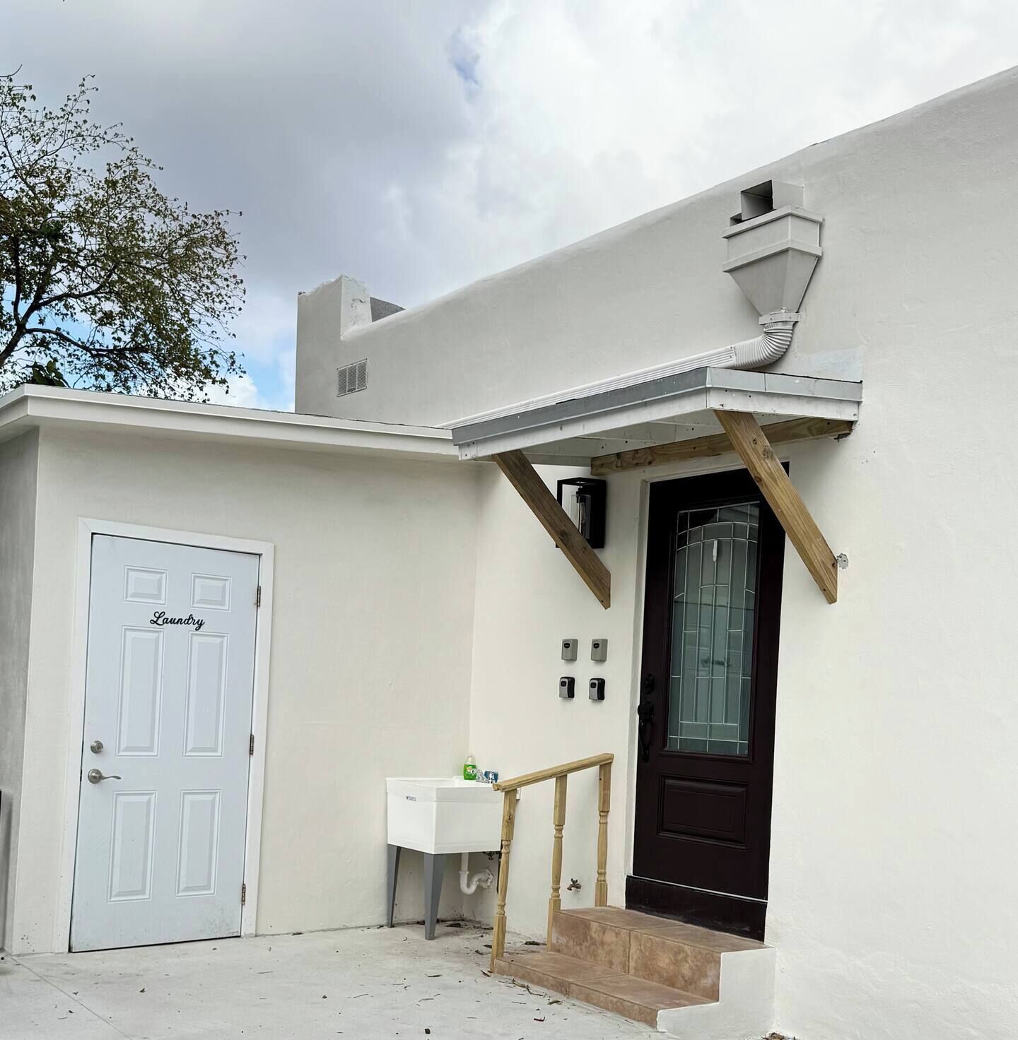 Un edificio bianco con una porta d'ingresso scura, un piccolo lavandino bianco e una scala in legno che conduce all'entrata.