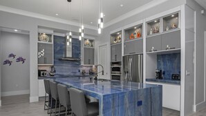 Una cocina moderna con una isla central, taburetes de bar, electrodomésticos integrados y objetos decorativos en estantes.