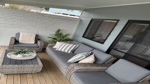 Un espace extérieur aménagé, abrité et doté de mobilier en osier et de coussins, une petite table avec une plante, et une vue sur la ville grâce à de grandes fenêtres.