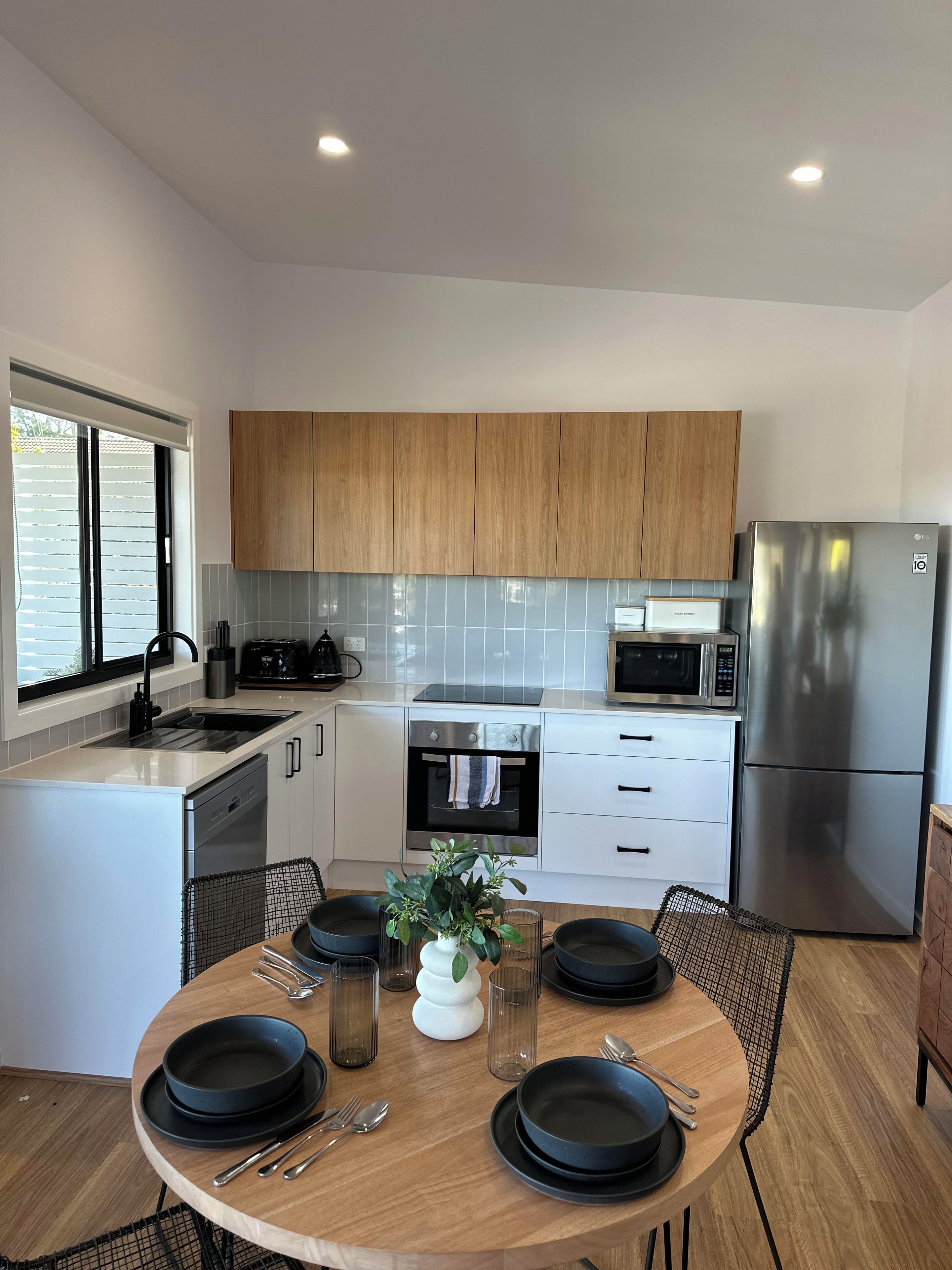A modern kitchen with wooden cabinets, stainless steel appliances, a dining area with a round table, and a window with blinds.