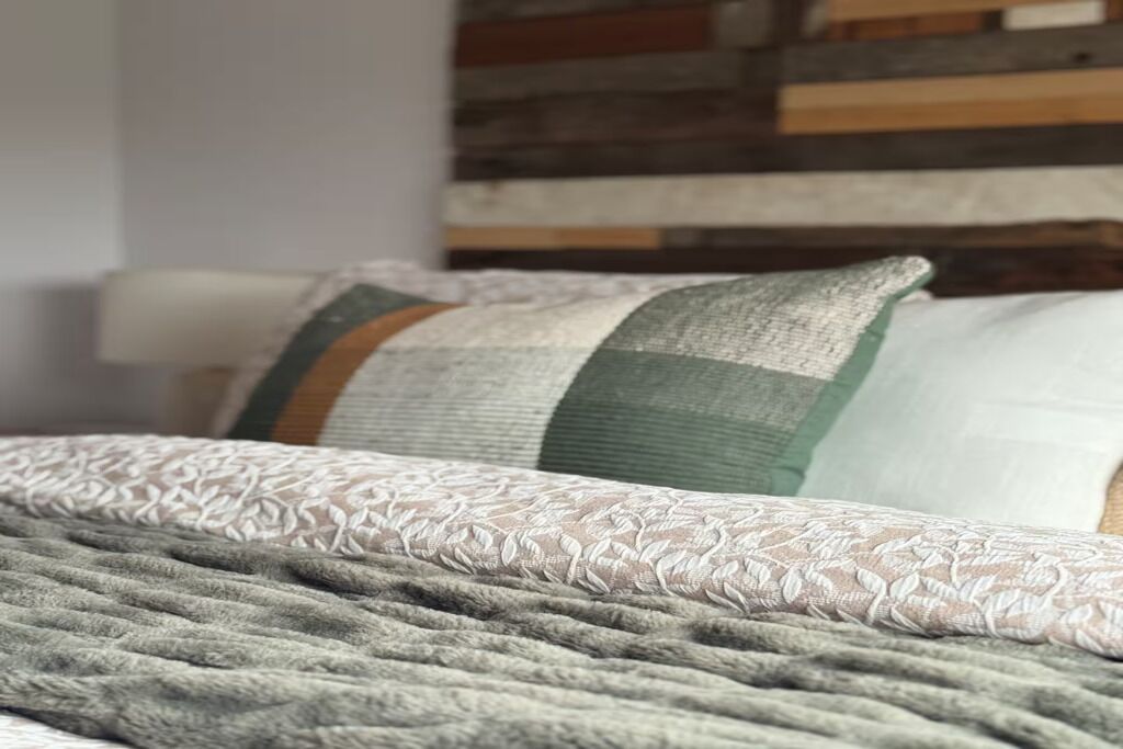 Ein Bett mit einer grauen Bettdecke und einem weißen Kopfkissen mit grün-weißer Musterung.