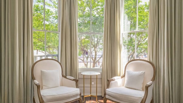 A room with two white upholstered chairs, a small round table, and a view of trees through large windows. - Mirabelle Suite 2 - Private Suite in Historic Savannah (Savannah)