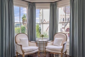 A corner room with two white upholstered chairs, a small round table with a vase, and large windows with grey curtains. - Mirabelle Suite 3 - Top Floor Suite in the Heart of Historic Savannah (Savannah)