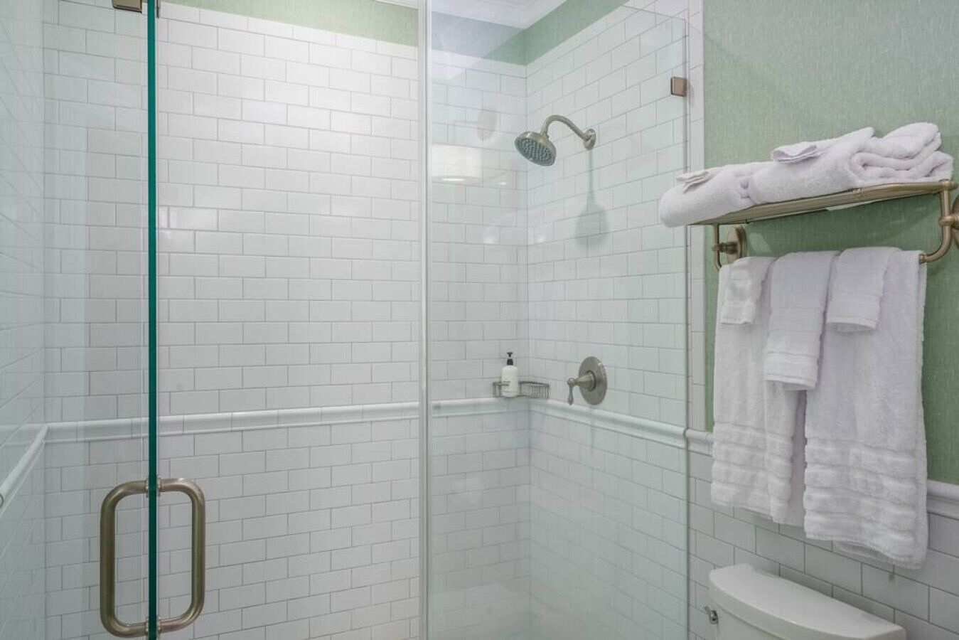 Ducha con cerramiento de vidrio, azulejos blancos, puerta de vidrio y toallero con toallas dobladas.