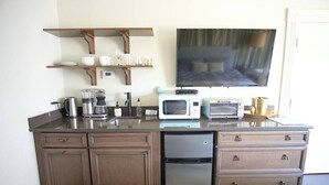 A compact kitchen with a microwave, toaster, coffee maker, and a sink. There are wooden cabinets and a flat-screen TV mounted on the wall.