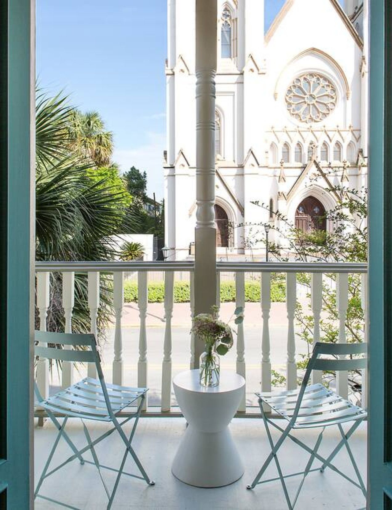 Un balcon avec deux chaises blanches et une table blanche ornée d’un vase de fleurs. On aperçoit, en arrière-plan, un bâtiment blanc doté d’une grande fenêtre circulaire et de fenêtres en arc de cercle.
