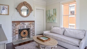 Un salón con chimenea de ladrillo, un espejo redondo, un sofá de color claro y una mesa de centro con un libro.