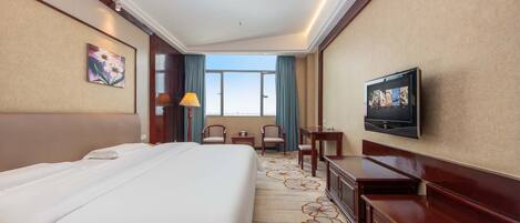 A hotel room with a large bed, a TV mounted on the wall, a desk with a chair, and a view of the sky through the window.