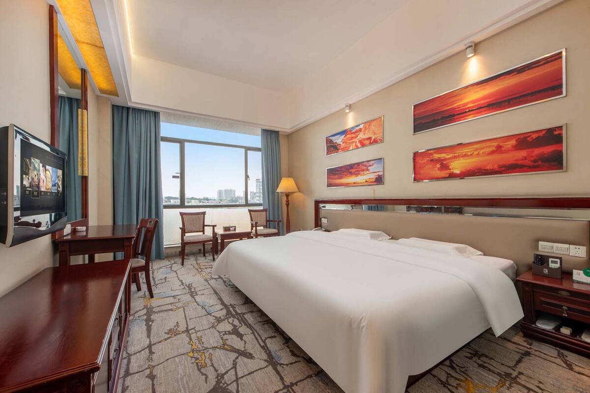 A hotel room with a large bed, a desk with a television, a chair, and a view of the city through large windows.