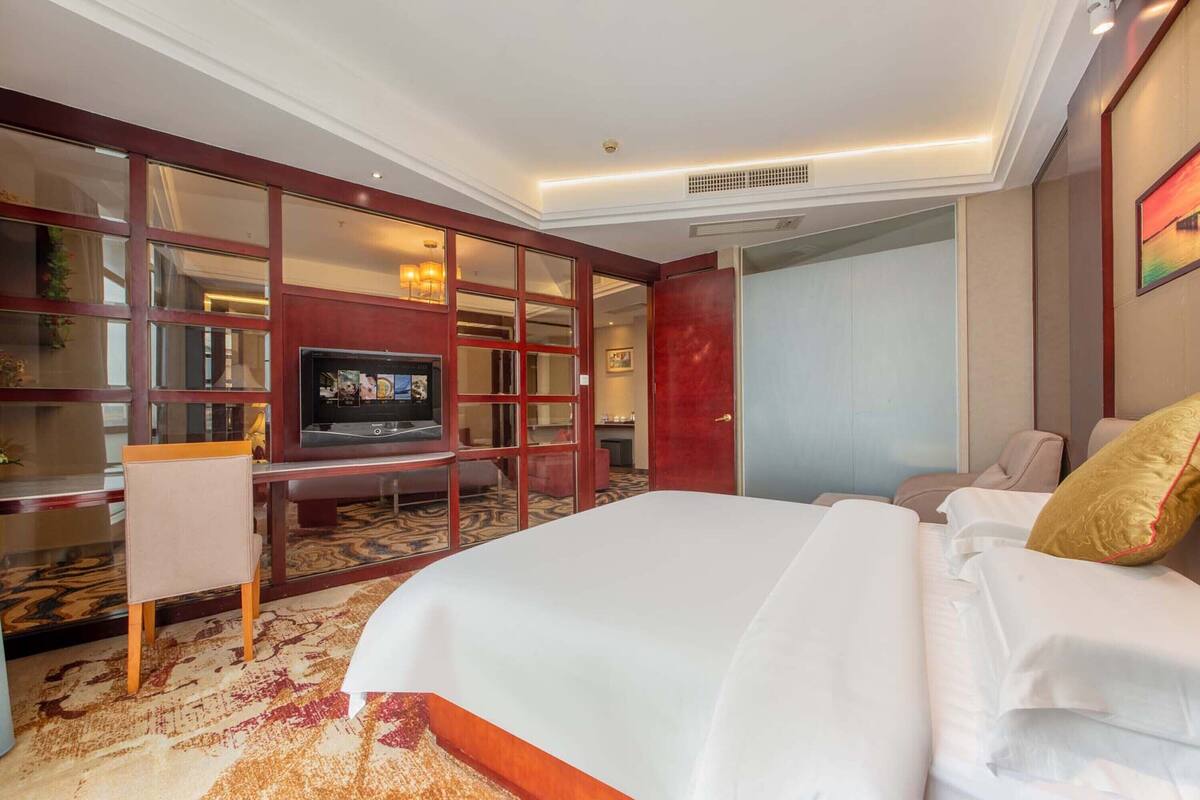 A hotel room with a large bed, a TV, a desk with a chair, and a bookshelf.