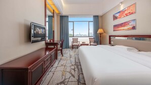 A hotel room with a large bed, a desk with a chair, a TV mounted on the wall, and a window with a city view. - Billions-Earn Hotel (Foshan)