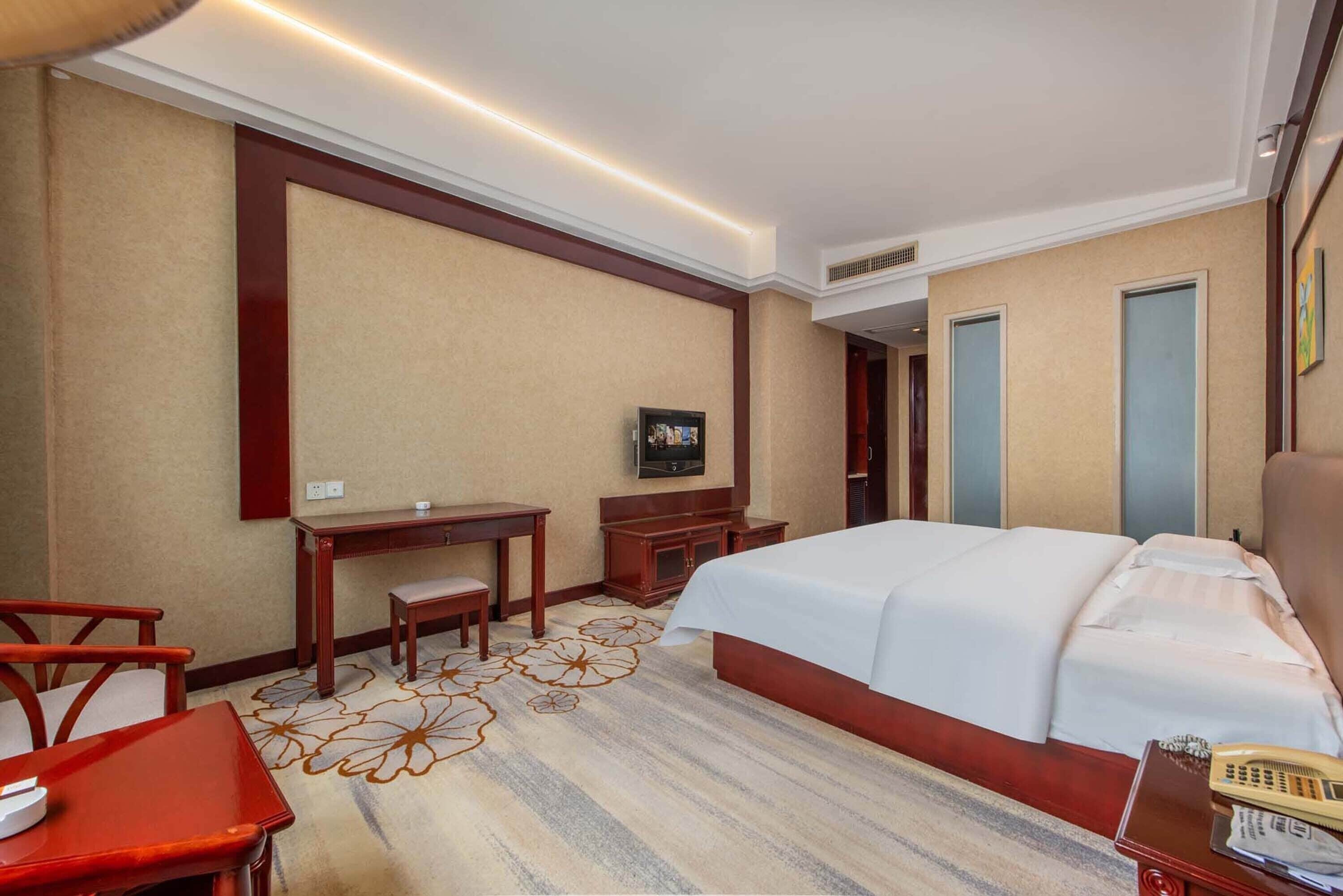 A hotel room with a large bed, a desk with a chair, a television, and a carpet with a floral pattern.