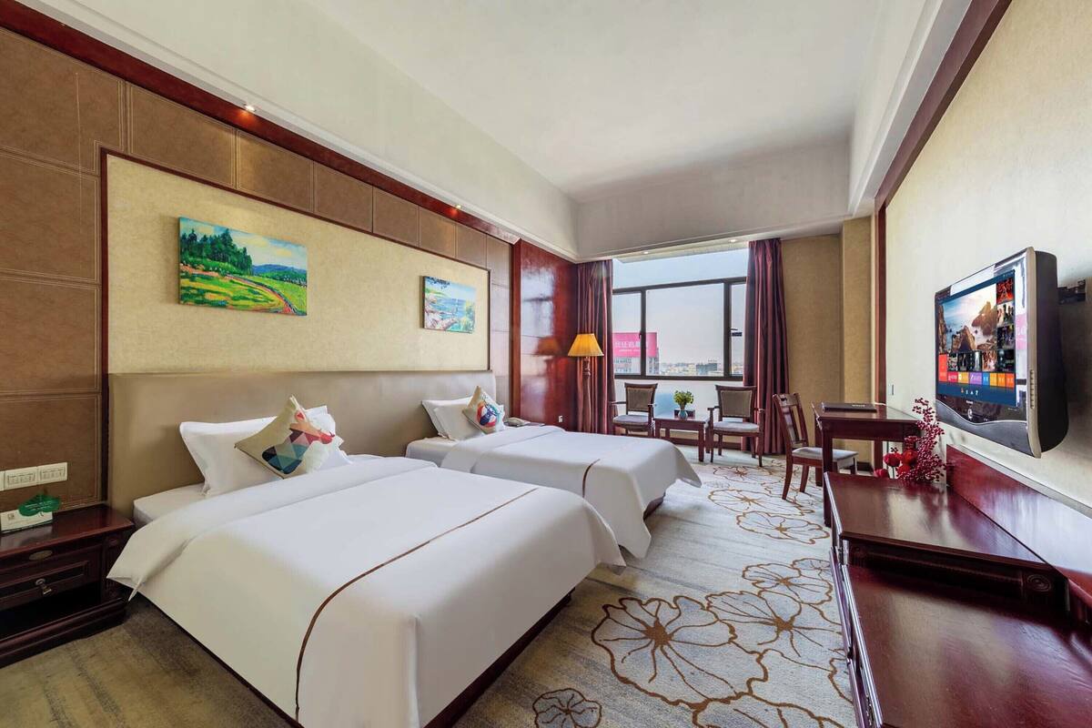 A hotel room with two beds, a TV, a desk, and a large window.