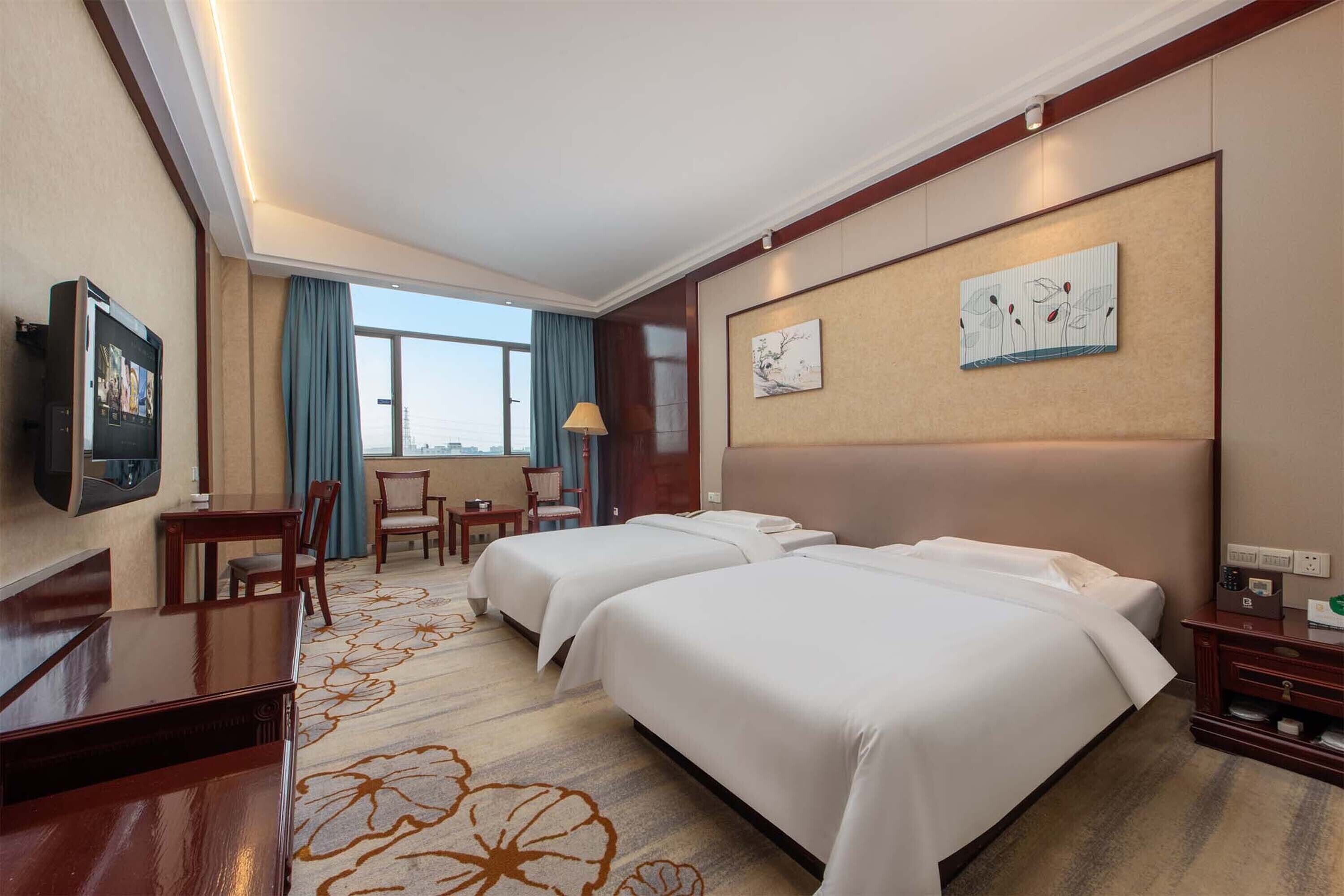 A hotel room with two beds, a desk, a chair, a TV, and a large window with a view.