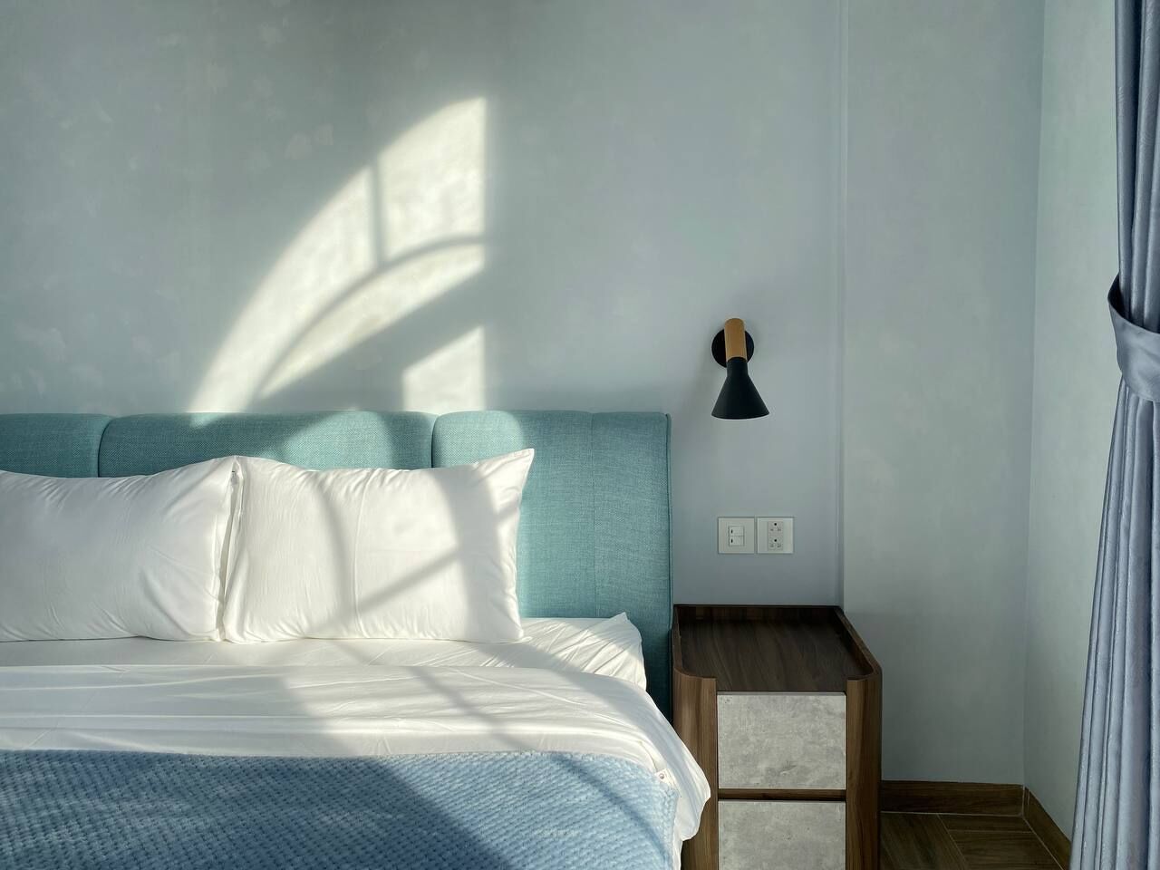 A neatly made bed with white linens and a teal headboard, a small wooden nightstand with a lamp, and a window casting shadows on the wall.