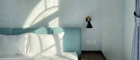 A neatly made bed with white linens and a teal headboard, a small wooden nightstand with a lamp, and a window casting shadows on the wall.