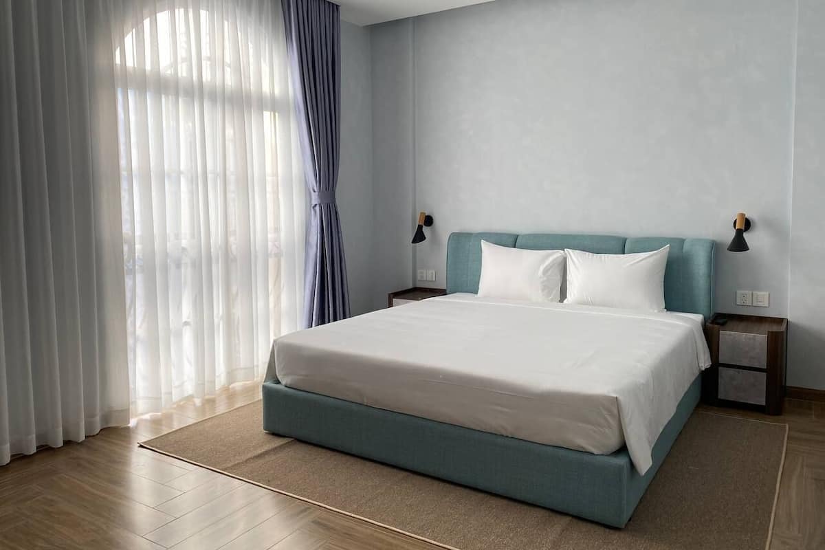 A modern hotel room with a large bed, light blue headboard, white bedding, and a view of the outdoors through sheer curtains.