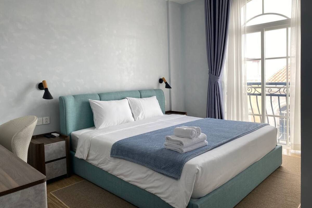 A neatly made bed with white and blue bedding, a bedside lamp, and a view of a balcony through a window with curtains.