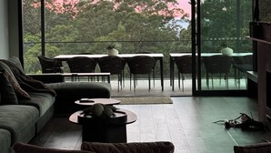 Ein modernes Wohnzimmer mit einem großen Glasfenster, das auf eine Terrassenausgang führt. Im Raum befinden sich eine Couch, ein Couchtisch und ein Essbereich mit Stühlen.