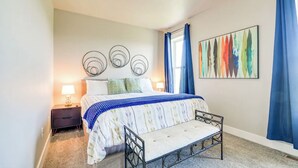 Een slaapkamer met een bed, nachtkastjes, een bankje, een kunstwerk aan de muur en een raam met gordijnen.