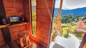 Ein Zimmer mit einem Laptop auf einem Regal, einem hölzernen Hocker und Blick auf eine gebirgige Landschaft durch ein großes Fenster.